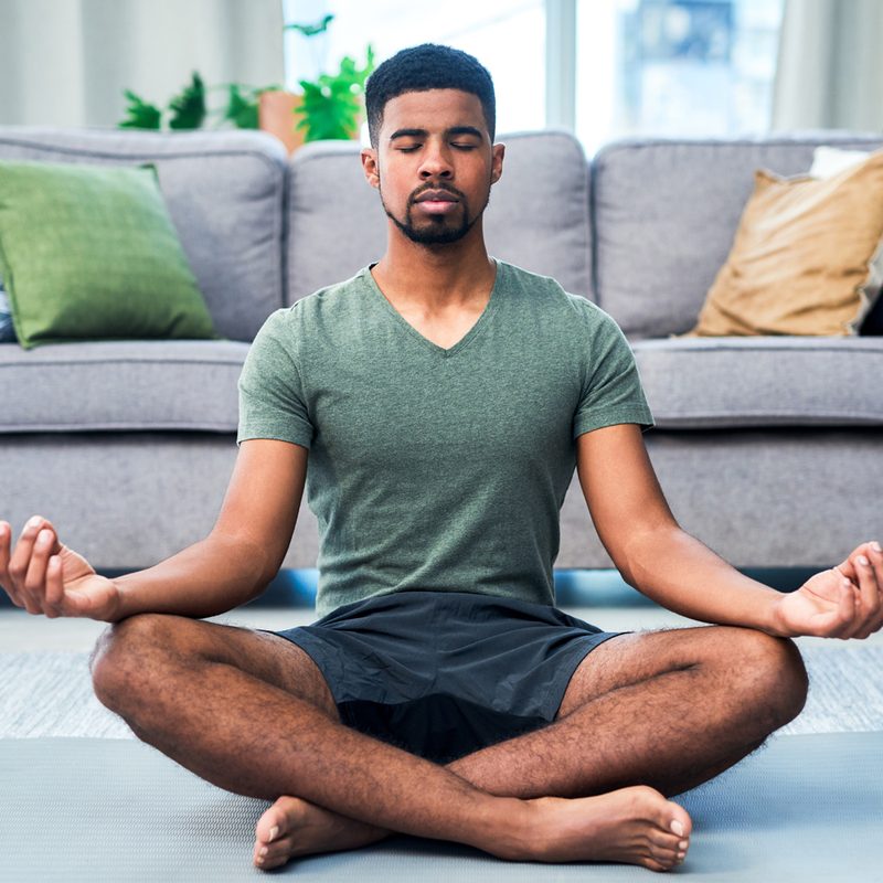 A man in a green shirt and gray shorts meditating on a yoga mat in the living room.