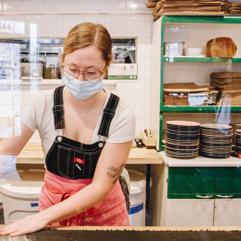 Sedona Kusler with facemask holding a cup as she wipes flour off the table in a kitchen.