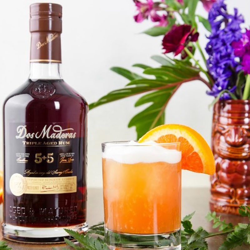 A glass of Pearl Diver cocktail garnished with orange beside a bottle of liquor and flowers along with plant stalks and a sliced orange