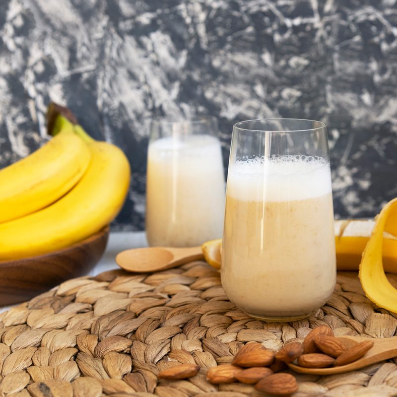 A banana shake with bananas, nuts, wooden spoons, coaster, and a glass of milk.