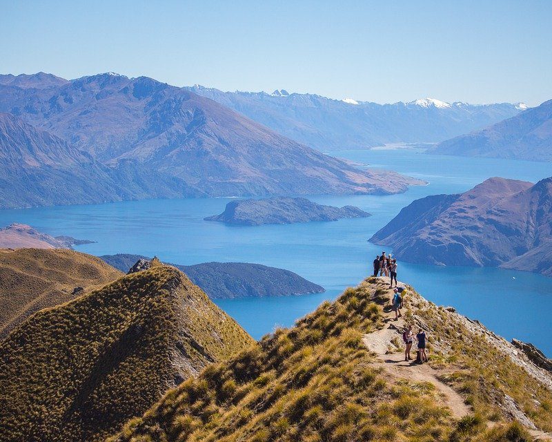 Several hikers on a large hill overlooking a big blue lake and several sourrounding mountains.