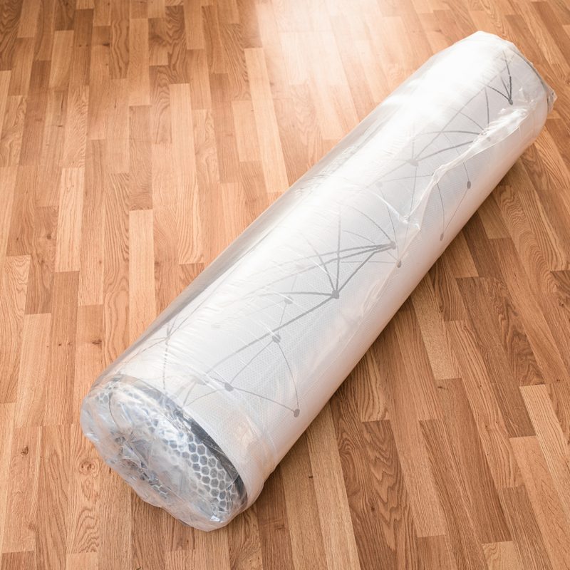 A rolled up latex mattress vacuum sealed in plastic on wooden floor.