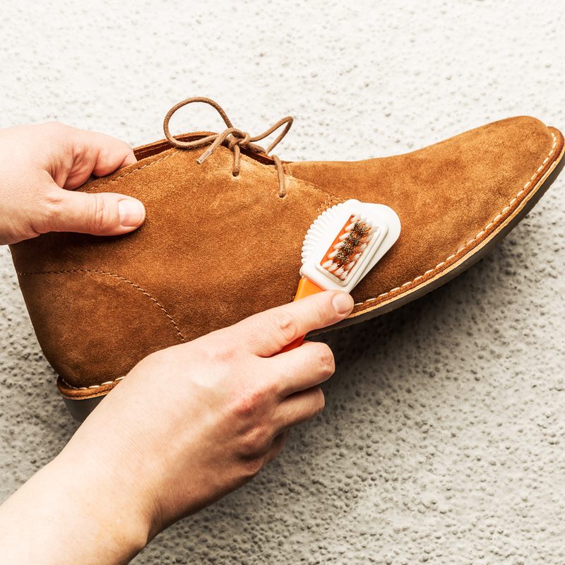 A pair of hands using a suede brush to clean a suede shoe on the floor.