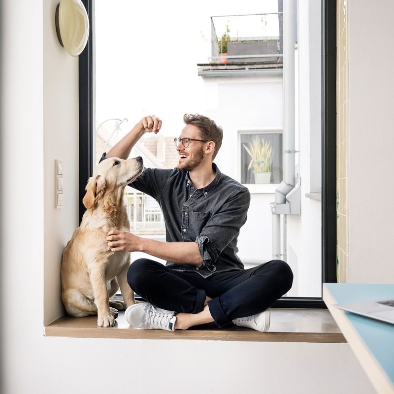 A bespectacled man in casual attire sitting beside his dog on the floor.