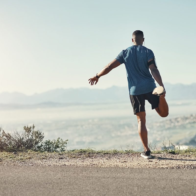 A man balancing on one leg to stretch on a road with a view of the mountain.