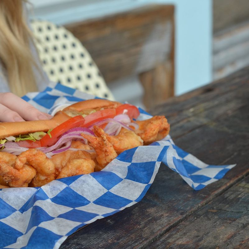 A close-up of a sandwich with shrimp, lemon, and veggies on a plate covered in blue and white paper beside a pitcher of water.