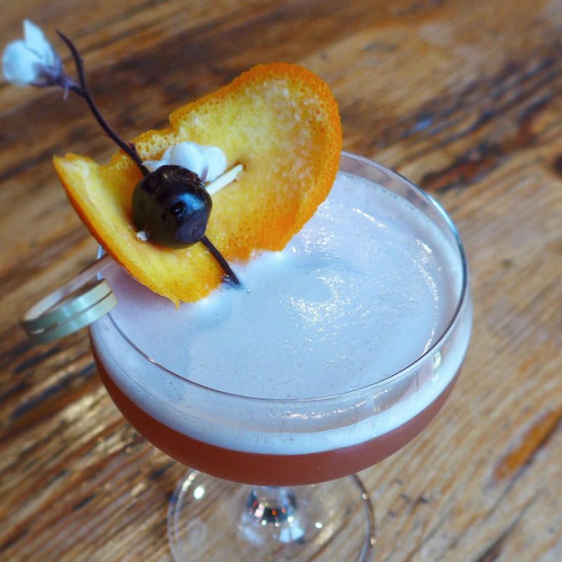 A glass of They Call Him Singapore "The Sling" garnished with orange on wooden table.