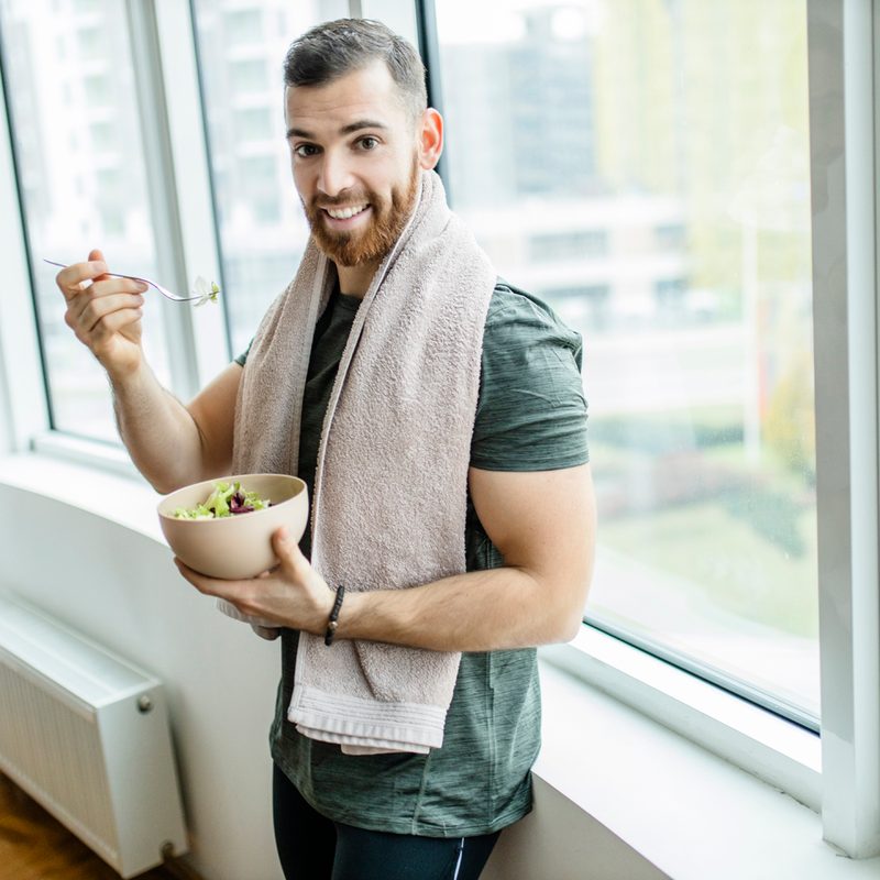 A man with a towel draped over his shoulder eating a salad after working out at home.