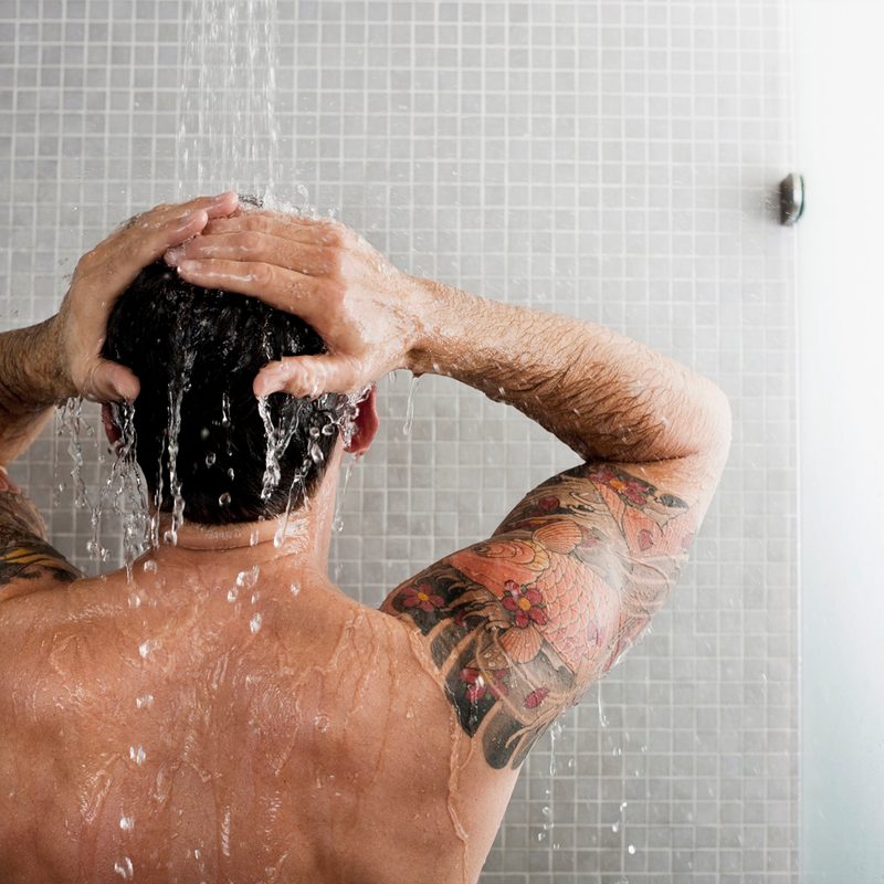 A man with tattoos on his biceps washing his hair in the shower.
