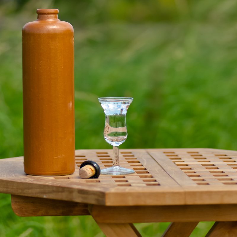 An opened bottle and a glass of genever beside a cork on a wooden round table with grass as background.