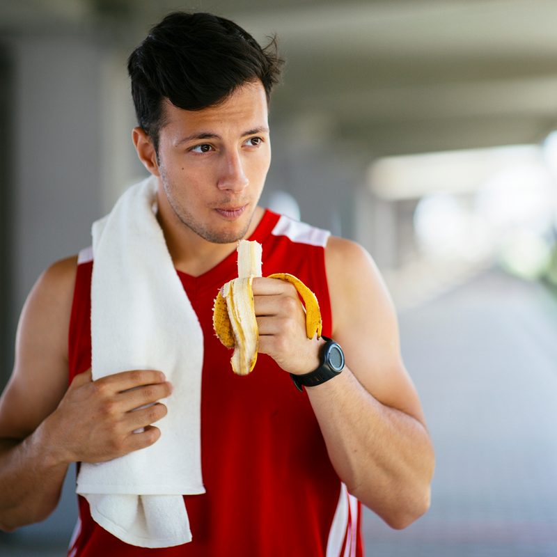 A man with a towel over his shoulder eating a banana.