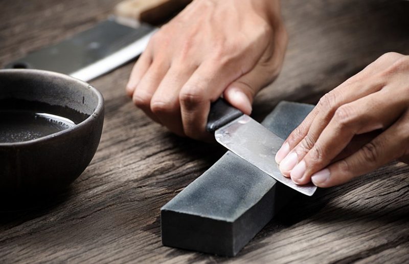 Hands sharpening a knife using a whetstone beside a bowl of water on a wooden table.