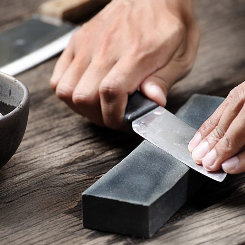 Hands sharpening a knife using a whetstone beside a bowl of water on a wooden table