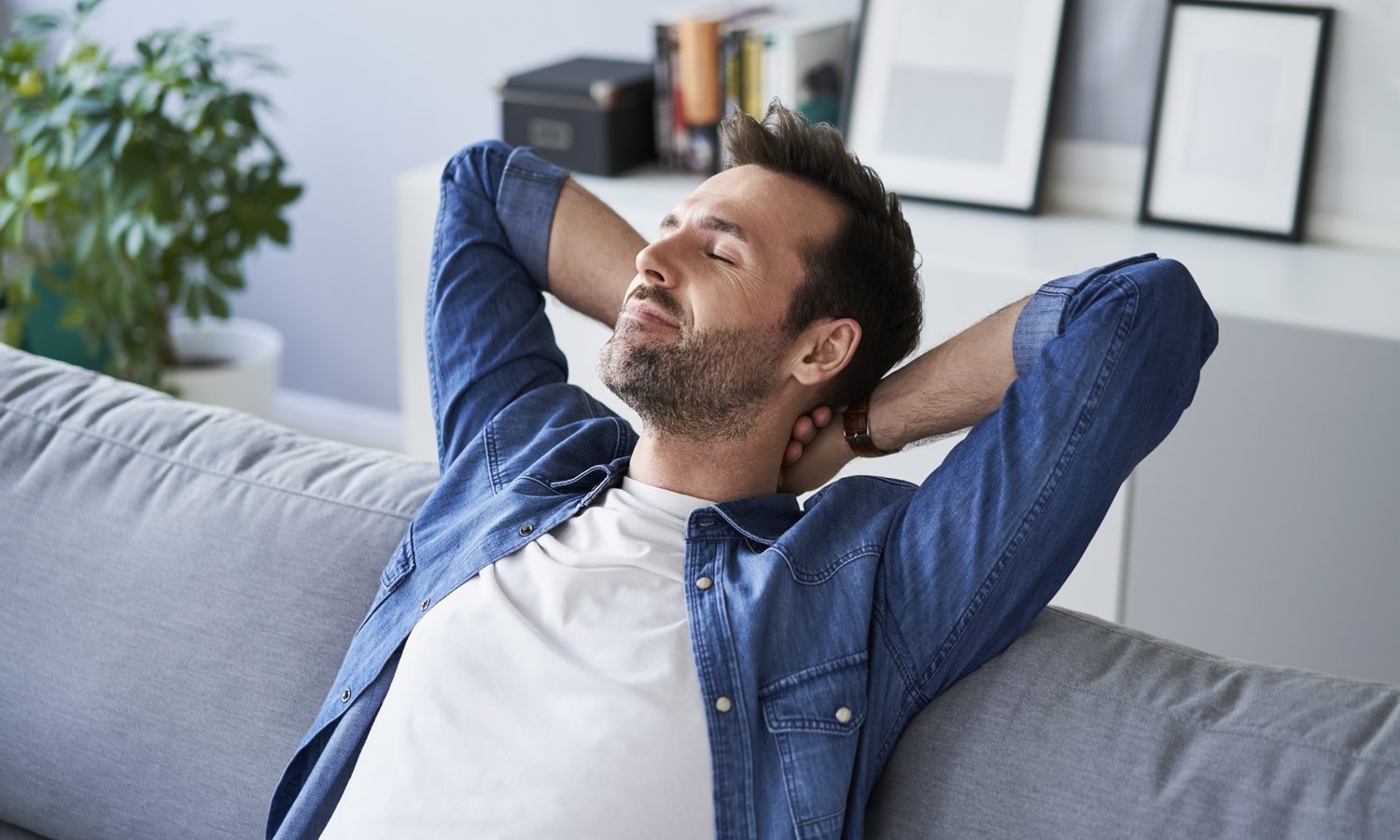 A man with his eyes closed resting on a gray sofa with his hands behind his neck.