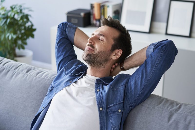 A man with his eyes closed resting on a gray sofa with his hands behind his neck.