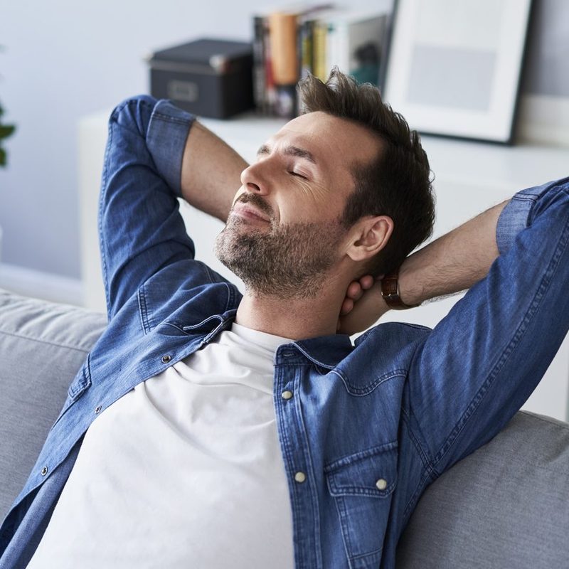A man with his eyes closed resting on a gray sofa with his hands behind his neck.