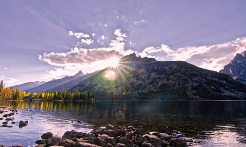 A Wyoming landscape, mountain lake and rocks leading up to jagged mountains with the sun setting behind them.