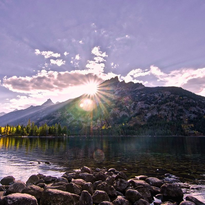 A Wyoming landscape, mountain lake and rocks leading up to jagged mountains with the sun setting behind them.