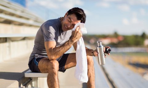 A man sitting on the bleachers holding a water bottle while wiping his brow with a white towel.
