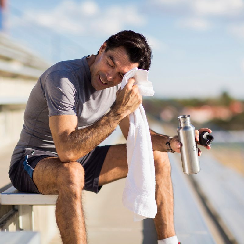 A man sitting on the bleachers holding a water bottle while wiping his brow with a white towel. 