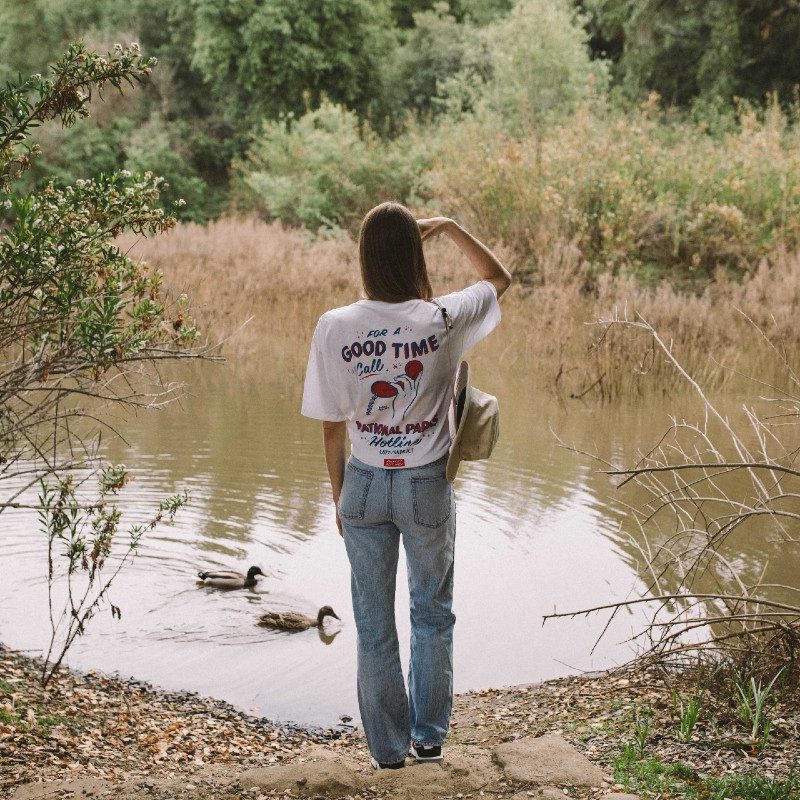 A model, seen from the back, wears a "For a Good Time Call Parks," Parks Project T-shirt while looking at ducks in a pond.