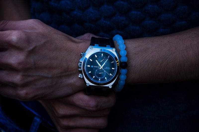 A man's wristwatch on a man's wrist alongside a blue-beaded bracelet.