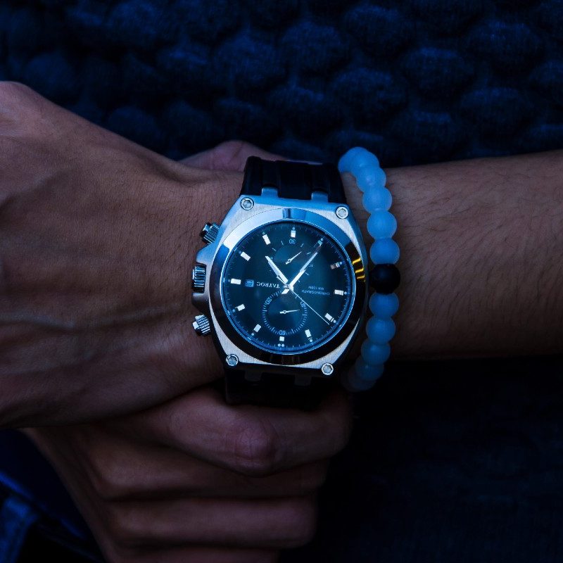 A man's wristwatch on a man's wrist alongside a blue-beaded bracelet.