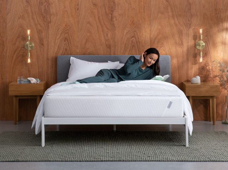A person reading on a Tuft and Needle Original Mattress in a room with a brown wall and gray flooring.