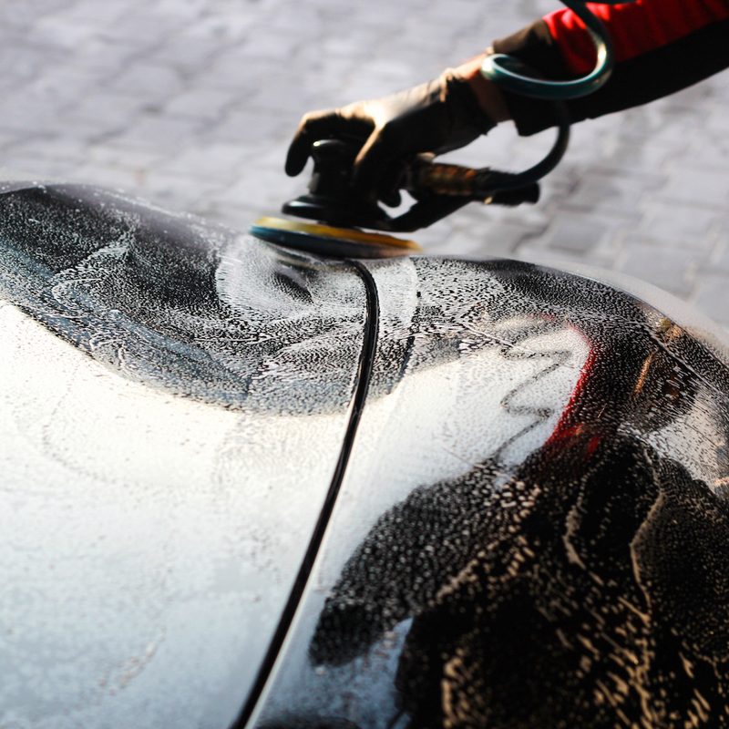 Man waxing a black car with a corded orbital buffer on a brick driveway.
