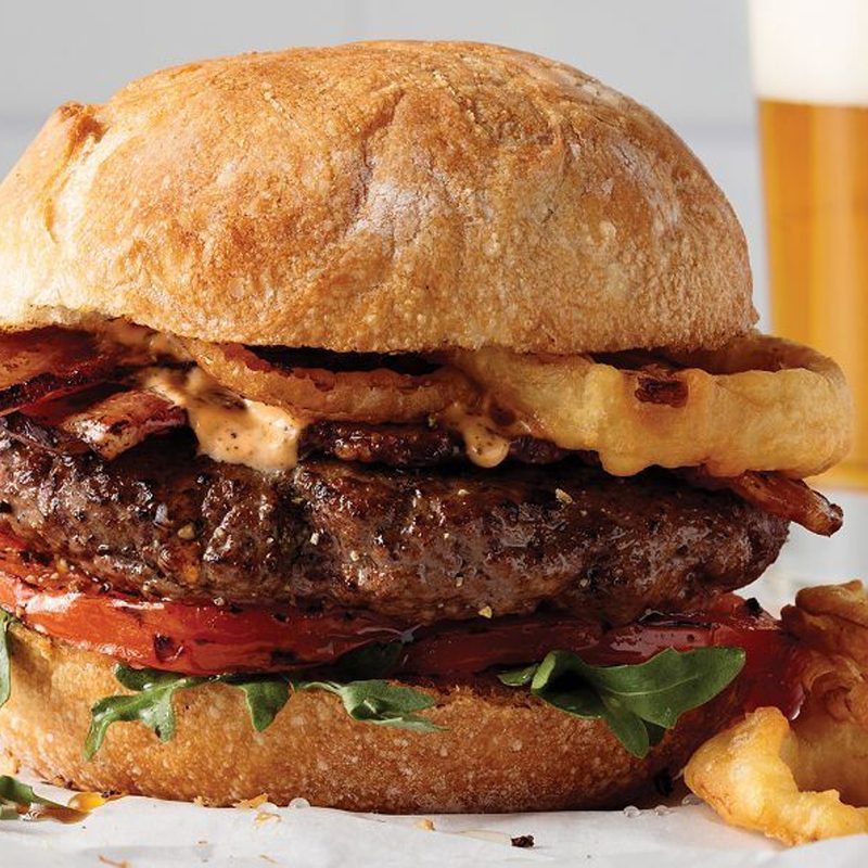 A burger with bacon and onion rings on it sits on a table next to a beer.