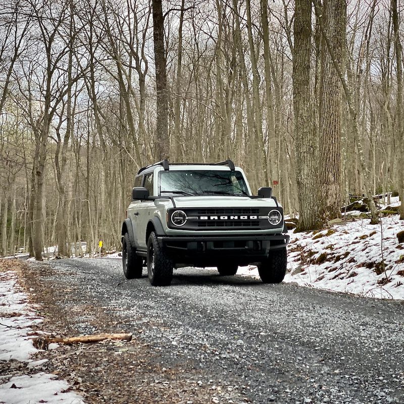 2021 Ford Bronco front end from passenger side angle on a gravel path in the woods with snow on the ground in front of bare trees