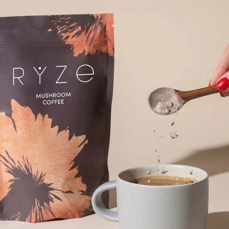 A hand dumps a dollop of Ryze Mushroom Coffee in a glass with a Ryze package behind it.