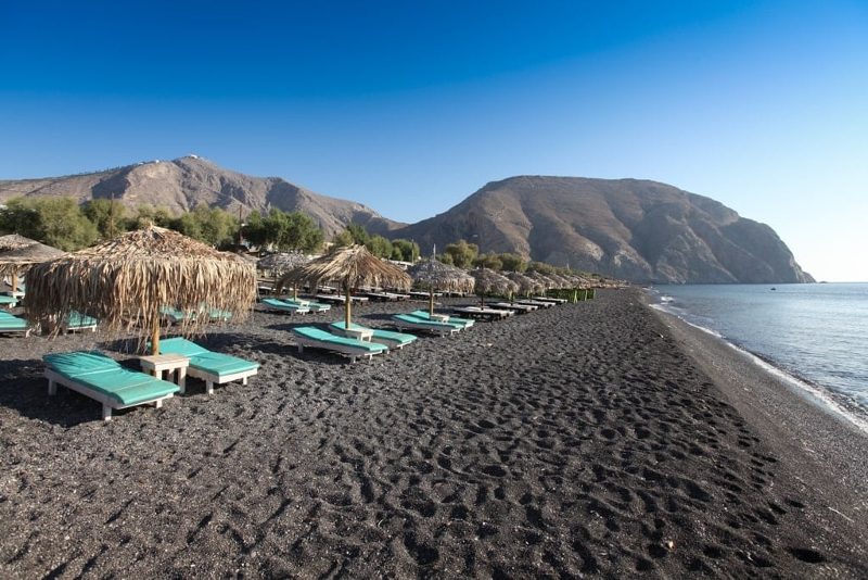 Lounge chairs lined up under thatched umbrellas on a black sand beach in Santorini.