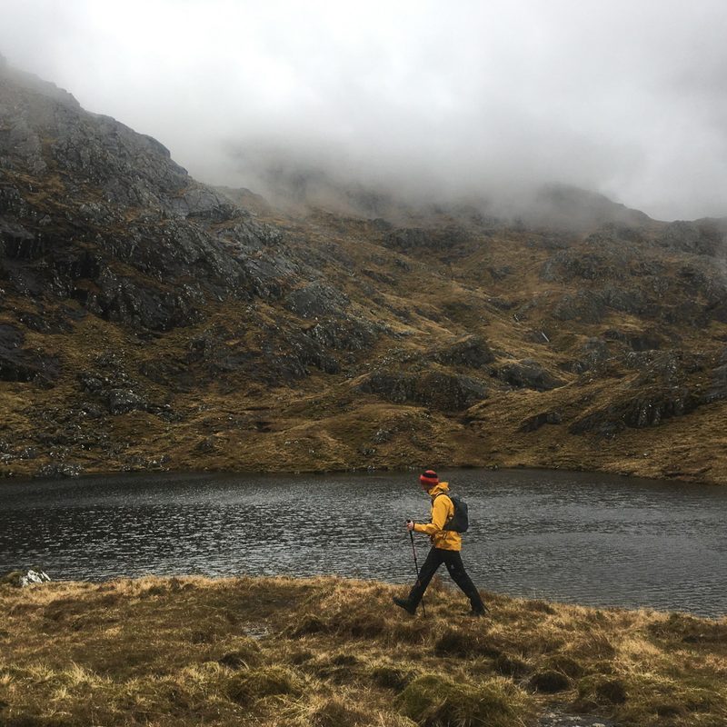 A man wearing a yellow jacket and red hat walks from right to left across a grassy mountain area. There is a small lake just behind him and the mountain tops are shrouded in clouds.