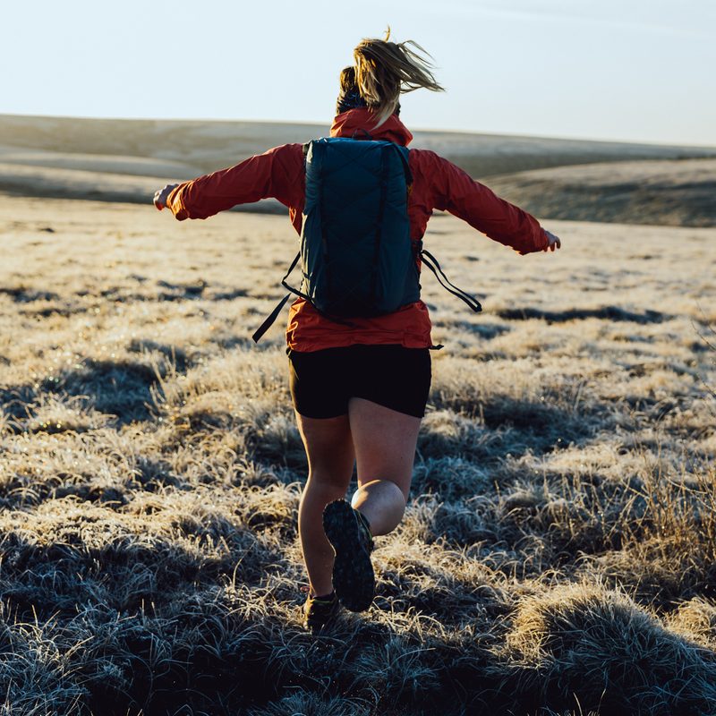 A woman runs away from the camera wearing a red jacket, shorts, and carrying a small blue backpack