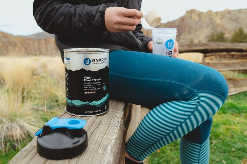 An outdoorsperson readies a plastic bottle for a Gnarly Vegan Protein powder supplement.