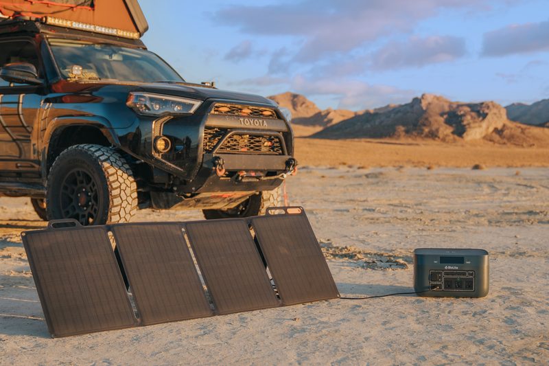 A dusty landscape with the front end of a 4x4 overland vehicle. A BioLite BaseCamp charging unit and solar system is set up in front of the truck.