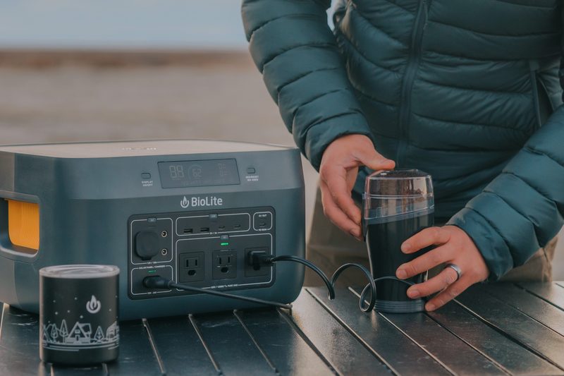 A close up image of a man in a down jacket using a coffee grinder that tis plugged into a BioLite BaseCharge unit.