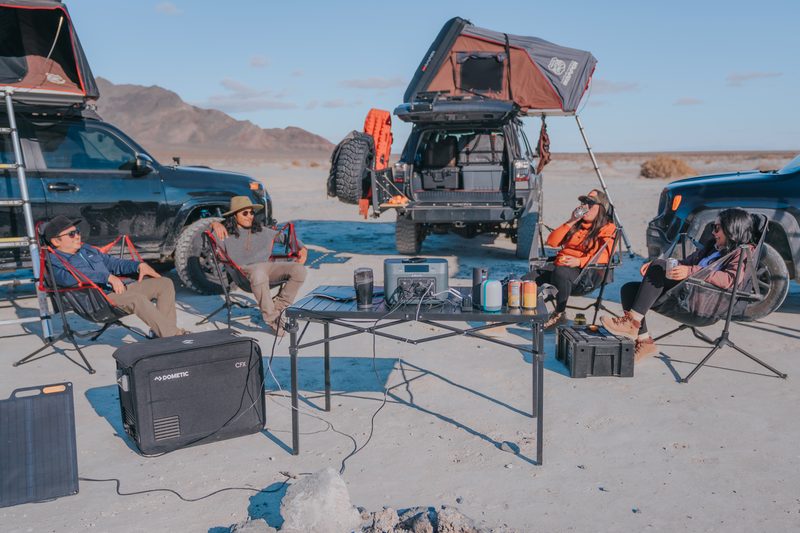 A backcountry camping setup with three off-road vehicles with roof tents. Four adults lounge in camping chairs with a table that has a BaseCharge power unit setup on it.