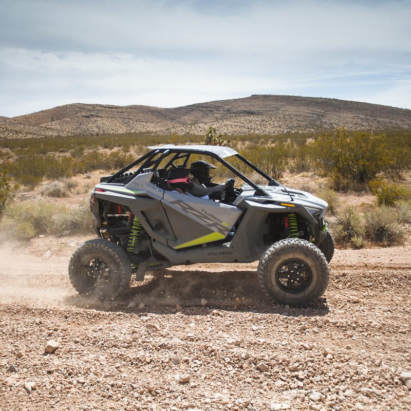 Closeup of side angle of Polaris RZR Turbo R on a dirt trail in the desert with mountains in the background.