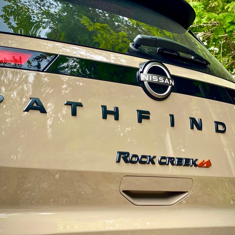 Close up of 2023 Nissan Pathfinder Rock Creek rear end badges in front of green trees.
