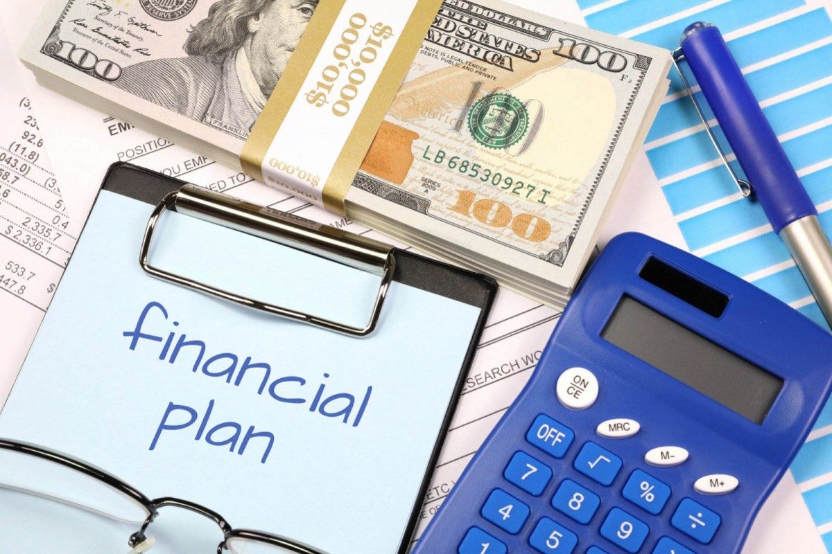 A clipboard with financial plan written on together with a bundle of dollar bills, accounts, a chart, a calculator, pen and glasses.