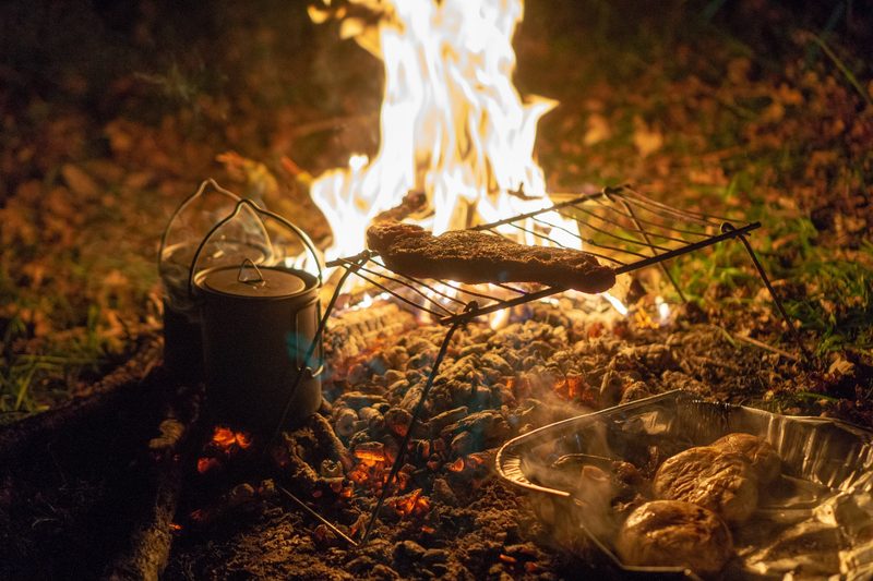 A steak cooks on a grill over some coals, while a campfire burns in the background