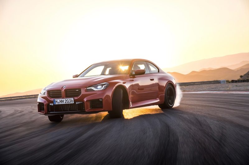 2023 BMW M2 drifting on a race track during a sunset and mountains in the back.