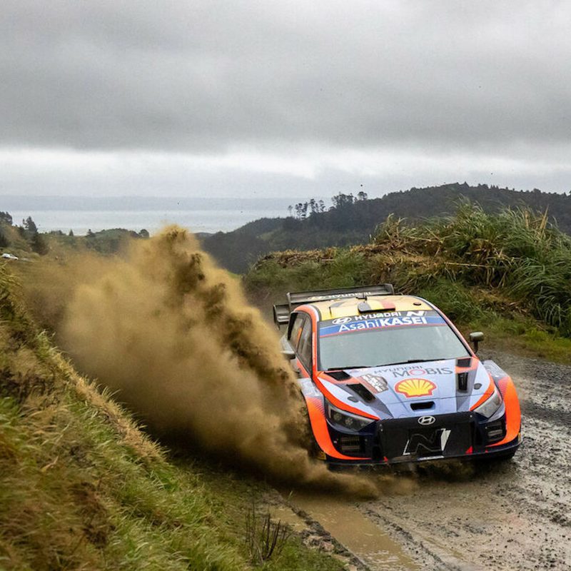 Hyundai i20N WRC rally car driving through thick mud on a dirt road with mountains in the back.