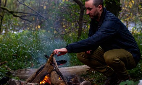 A man puts another log on the campfire wearing Ibex Nomad pants.