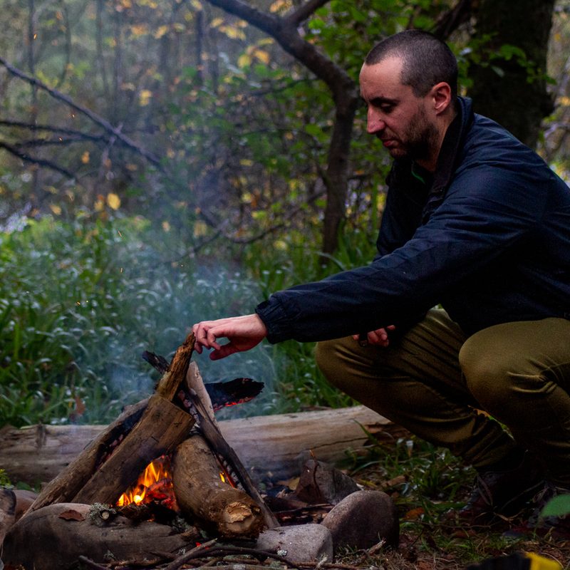 A man puts another log on the campfire wearing Ibex Nomad pants.