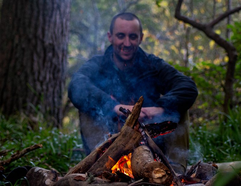 A man sits on the far side of the campfire while smoke rises in front of him.
