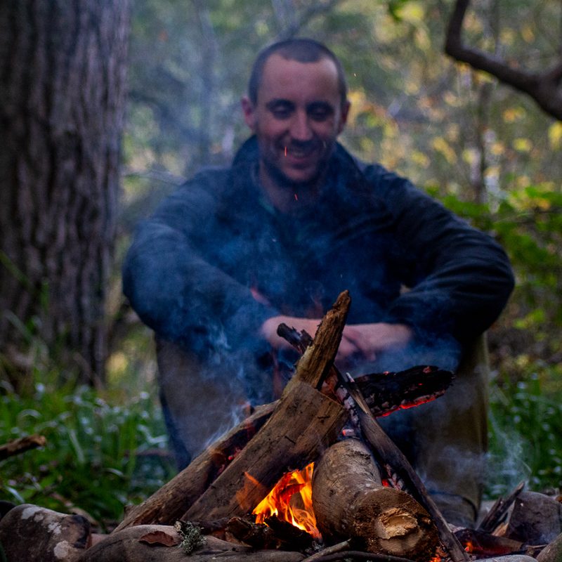 A man sits on the far side of the campfire while smoke rises in front of him.