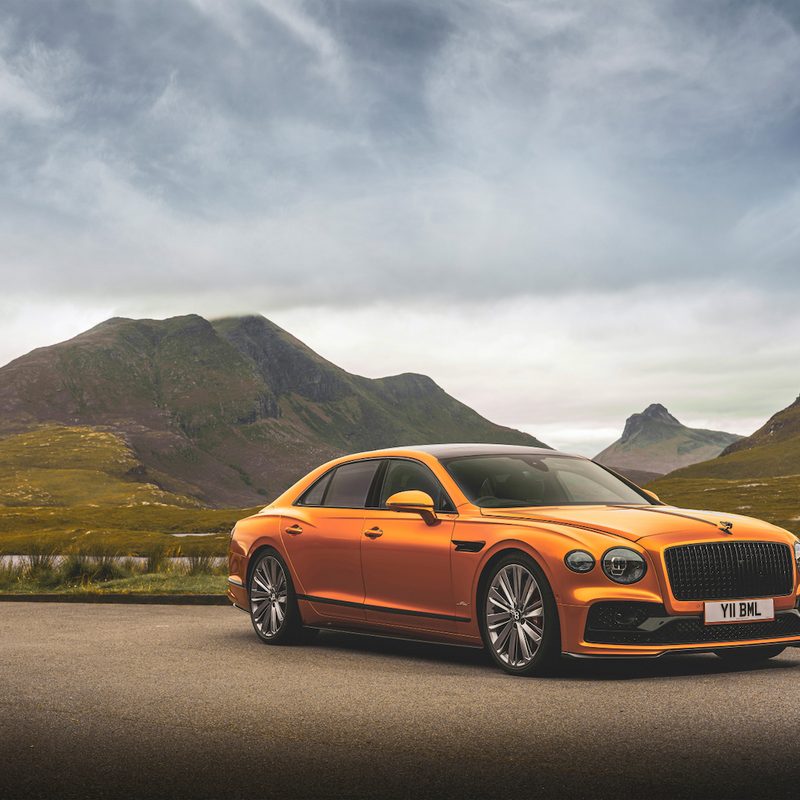 Orange 2022 Bentley Flying Spur parked in front of mountains with cloud skies in the back.